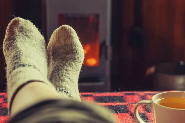 Feet legs in winter clothes wool socks and cup tea at fireplace background. Woman sitting at home on winter or autumn evening relaxing and warming up. Winter and cold weather concept. Hygge Christmas eve