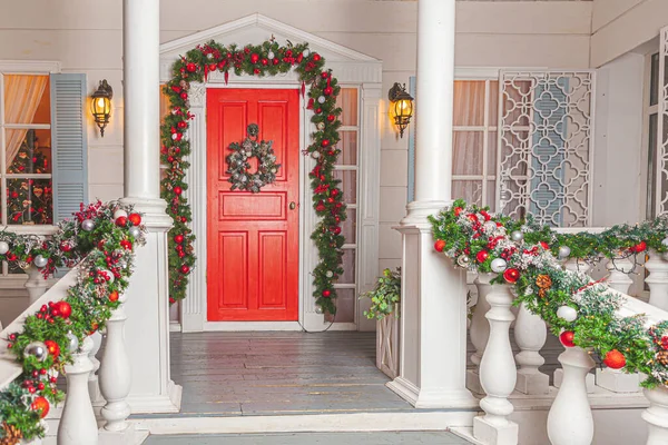 Christmas porch decoration idea. House entrance with red door decorated for holidays. Red and green wreath garland of fir tree branches and lights on railing. Christmas eve at home.