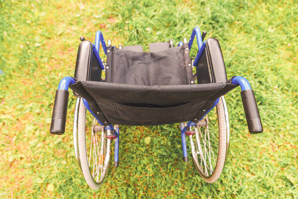 Empty wheelchair standing on grass in hospital park waiting for patient services. Invalid chair for disabled people parked outdoor in nature. Handicap accessible symbol. Health care medical concept