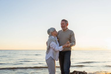 Yürümeye devam. Romantik olgun çift plaj gezisinde birlikte dans ediyor. Mutlu gülümseyen aile emeklisi kadın koca eş birlikte eğleniyor. Aile zamanı aşk bakımı