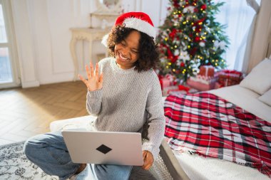 Mutlu noeller. Noel Baba şapkalı Afrikalı kadın bilgisayarında görüntülü konuşma yapıyor. Sosyal ağda neşeli ruh hali kayıt videosu blogcusu. Noel 'i kutlamak için arkadaşlarıyla görüntülü görüşme yapan bir kız.