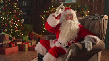 Mutlu noeller. Mutlu Noel Baba şenlikli oturma odasında Noel ağacı süslemeli sandalyede oturuyor. Noel arifesinde evde Noel Baba. Kış Yeni Yıl Tatili Kutlamaları