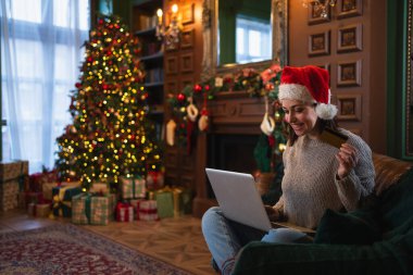 Mutlu noeller. Bilgisayarla alışveriş yapan genç bir kadın Noel hediyeleri için altın kredi kartıyla ödeme yapıyor. İnternet üzerinden alışveriş yapan kız, kredi kartı bilgilerine evde kış tatilinin tadını çıkarıyor.