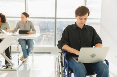 Erişilebilir çevre. Modern ofiste dizüstü bilgisayarla çalışan engelliler için sandalyedeki genç adam. Birlikte çalışan çok ırklı bir grup insan. Genç profesyonel takım