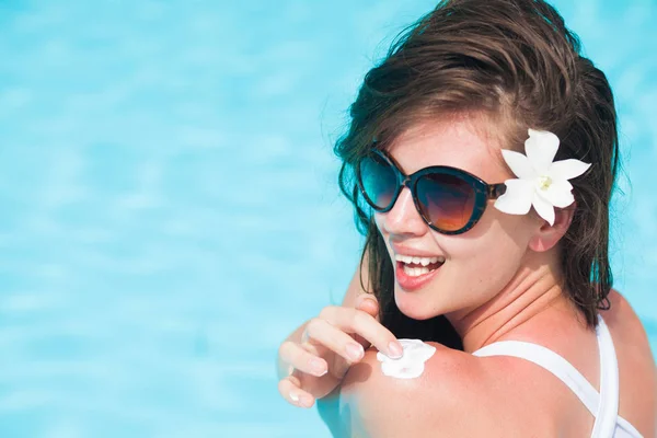 Jovem mulher de cabelos compridos em óculos de sol e com flor colocando creme solar no ombro — Fotografia de Stock