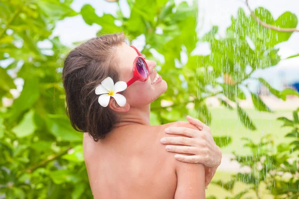 Mujer joven con flor de frangipany en el pelo refrescante en la ducha tropical —  Fotos de Stock