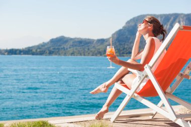 Genç kadının şezlongda dinlenip Lago di Garda'da Aperol Spritz kokteyli içen portresi
