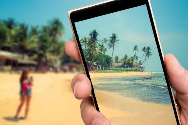 Akıllı telefon ekranında okyanus kıyısındaki kumsal. Tropiklerde yaz güneşli bir gün. Sahilde palmiye ağaçları.