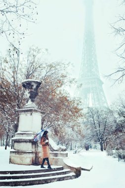 Bej ceketli genç bir kadın, Eyfel Kulesi'nin arka planında karlı bir kış Paris'te şemsiyenin altında yürüyor. Romantik esmer kız Paris'te düşen kar arka planda yürüyor. Karlı paris
