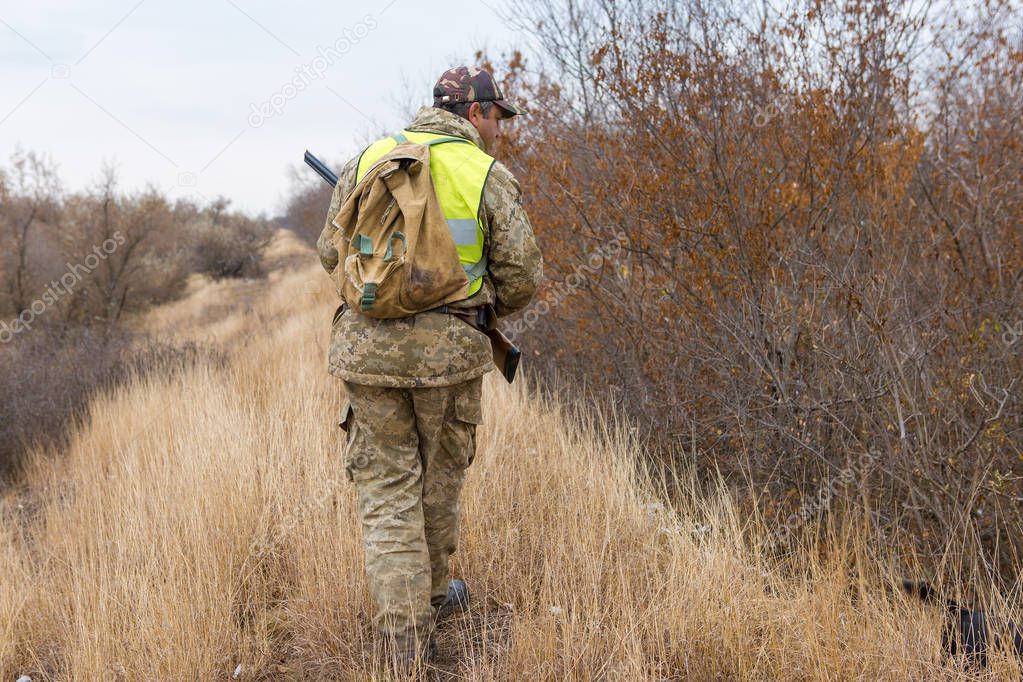 A hunter in a hat with a gun in camouflage and a reflective vest in the steppe with dog