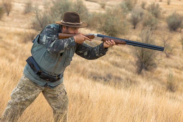 Bearded hunter in a hat stands with a gun and looks into the distance in search of prey.