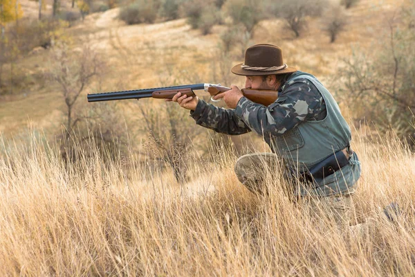Bearded hunter in a hat stands with a gun and looks into the distance in search of prey.