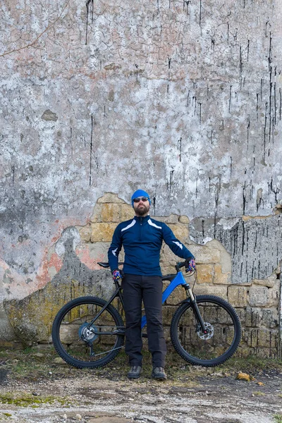 A guy in sportswear riding clothes on a modern mountain carbon bike with an air suspension fork at a vintage brick concrete wall.