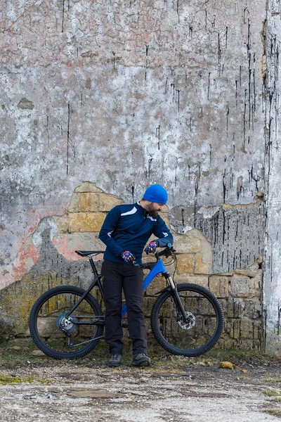 A guy in sportswear riding clothes on a modern mountain carbon bike with an air suspension fork at a vintage brick concrete wall.