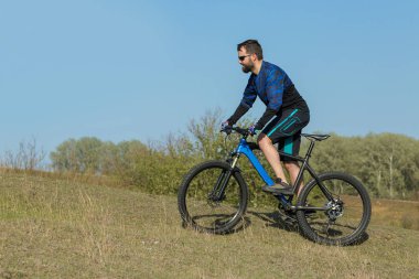 Bisikletçi şort ve mayo modern bir karbon hardtail bisiklet ile bir hava süspansiyon çatal sürmek off-road ormana yakın yeşil tepelerde