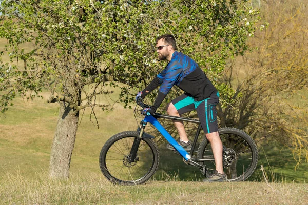 Cyclist in shorts and jersey on a modern carbon hardtail bike with an air suspension fork rides off-road on green hills near the forest