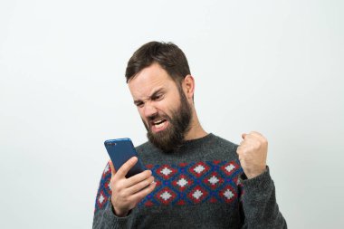 Angry young bearded caucasian man watching smartphone on white background