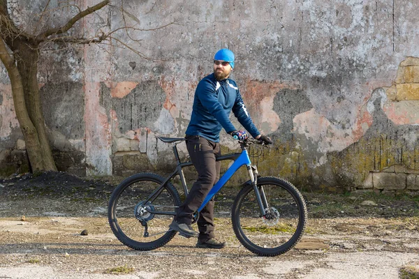 A guy in sportswear riding clothes on a modern mountain carbon bike with an air suspension fork at a vintage brick concrete wall.