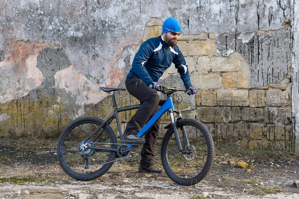 A guy in sportswear riding clothes on a modern mountain carbon bike with an air suspension fork at a vintage brick concrete wall.