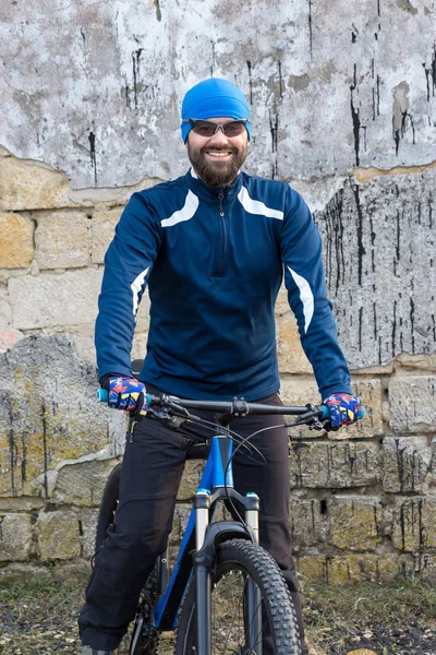 A guy in sportswear riding clothes on a modern mountain carbon bike with an air suspension fork at a vintage brick concrete wall.