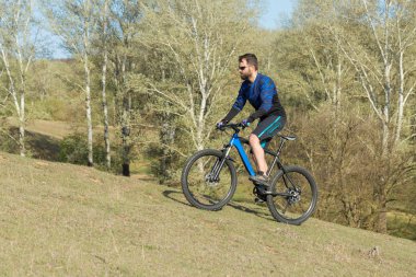 Bisikletçi şort ve mayo modern bir karbon hardtail bisiklet ile bir hava süspansiyon çatal sürmek off-road ormana yakın yeşil tepelerde