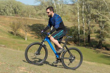 Bisikletçi şort ve mayo modern bir karbon hardtail bisiklet ile bir hava süspansiyon çatal sürmek off-road ormana yakın yeşil tepelerde