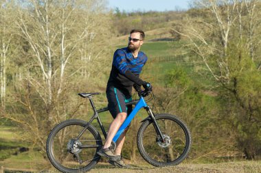 Bisikletçi şort ve mayo modern bir karbon hardtail bisiklet ile bir hava süspansiyon çatal sürmek off-road ormana yakın yeşil tepelerde