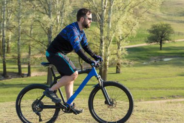Bisikletçi şort ve mayo modern bir karbon hardtail bisiklet ile bir hava süspansiyon çatal sürmek off-road ormana yakın yeşil tepelerde