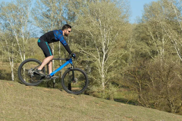 Cyclist in shorts and jersey on a modern carbon hardtail bike with an air suspension fork rides off-road on green hills near the forest