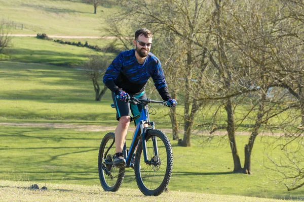 Cyclist in shorts and jersey on a modern carbon hardtail bike with an air suspension fork rides off-road on green hills near the forest