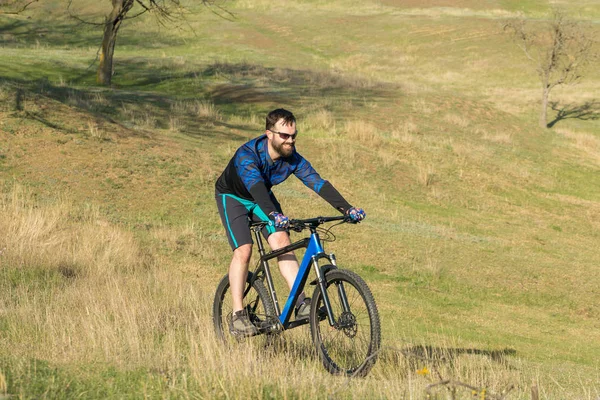 Cyclist in shorts and jersey on a modern carbon hardtail bike with an air suspension fork rides off-road on green hills near the forest