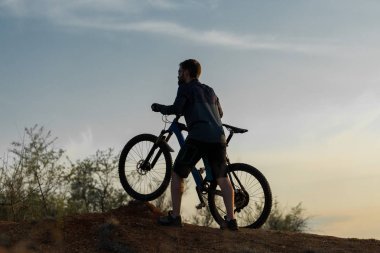 Şort ve mayo modern bir karbon hardtail bisiklet üzerinde bir hava süspansiyon çatal sürmek off-road turuncu-kırmızı tepelerde yaz aylarında gün batımı akşamı