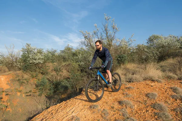 Cyclist in shorts and jersey on a modern carbon hardtail bike with an air suspension fork rides off-road on the orange-red hills at sunset evening in summer