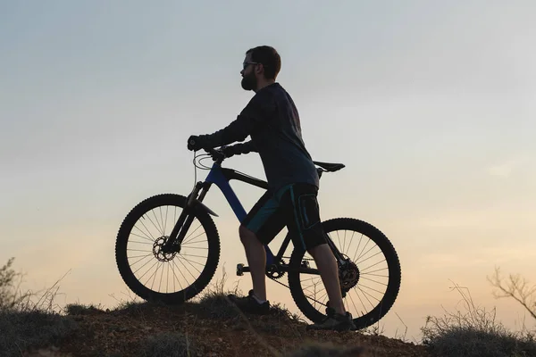 Cyclist in shorts and jersey on a modern carbon hardtail bike with an air suspension fork rides off-road on the orange-red hills at sunset evening in summer