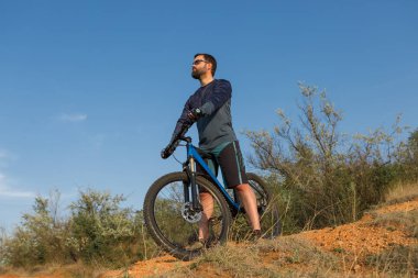 Taze yeşil yaz ormanının arka planına karşı bir uçurumun üzerinde duran bir hava süspansiyon çatal ile modern bir karbon hardtail bisiklet üzerinde şort ve mayo Bisikletçi