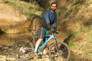 Taze yeşil yaz ormanının arka planına karşı bir uçurumun üzerinde duran bir hava süspansiyon çatal ile modern bir karbon hardtail bisiklet üzerinde şort ve mayo Bisikletçi