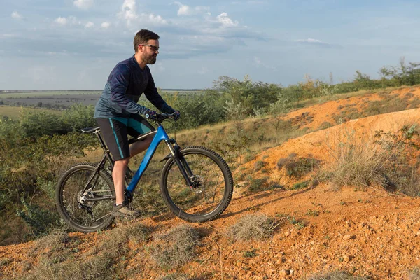 Cyclist in shorts and jersey on a modern carbon hardtail bike with an air suspension fork standing on a cliff against the background of fresh green summer forest