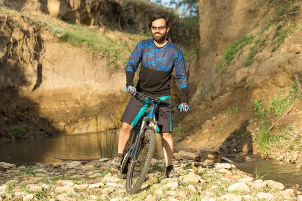 Cyclist in shorts and jersey on a modern carbon hardtail bike with an air suspension fork standing on a cliff against the background of fresh green summer forest