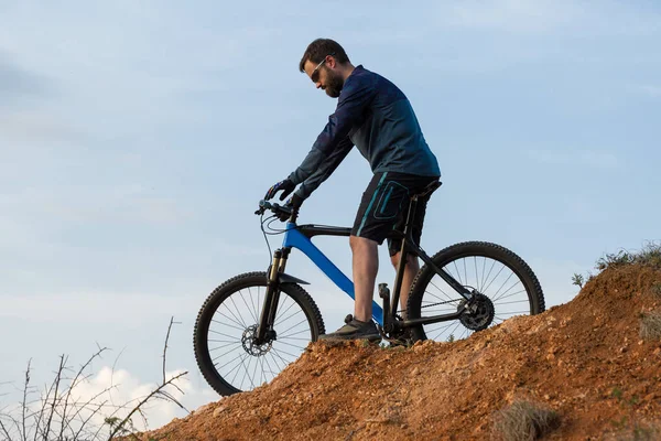 Cyclist in shorts and jersey on a modern carbon hardtail bike with an air suspension fork rides off-road on the orange-red hills at sunset evening in summer