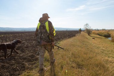 Sırt çantalı bir avcı ve arka planda bir av tüfeği.