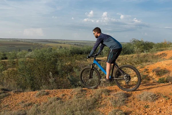 Cyclist in shorts and jersey on a modern carbon hardtail bike with an air suspension fork rides off-road on the orange-red hills at sunset evening in summer