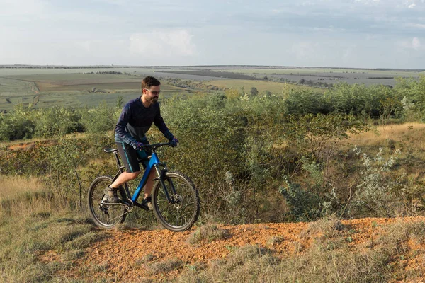 Cyclist in shorts and jersey on a modern carbon hardtail bike with an air suspension fork rides off-road on the orange-red hills at sunset evening in summer