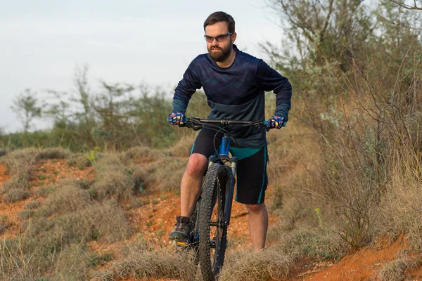 Cyclist in shorts and jersey on a modern carbon hardtail bike with an air suspension fork rides off-road on the orange-red hills at sunset evening in summer