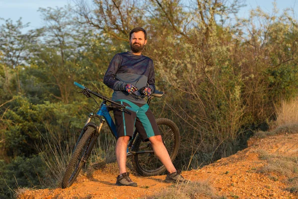 Cyclist in shorts and jersey on a modern carbon hardtail bike with an air suspension fork rides off-road on the orange-red hills at sunset evening in summer