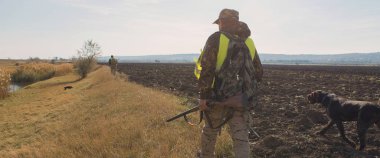 Silahlı bir avcı ve bir köpek bozkırda ilk kara çıkarlar yansıtıcı yelek giymiş sülün avlarlar.
