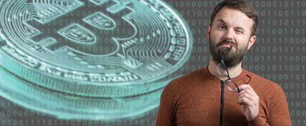 A pensive guy with a beard and glasses against a background of Bitcoin. Thinking about question, pensive expression, looks incredulous. Change in the growth and fall of cryptocurrency.