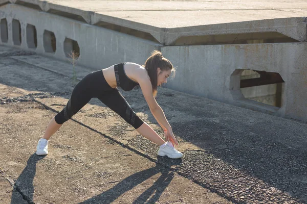 İnce atletik kız bitmemiş bir binanın çatısında germe egzersizleri gerçekleştirir, kentsel arka plan.
