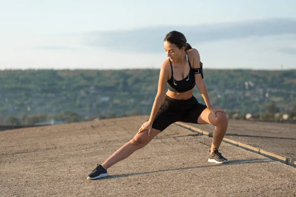 İnce atletik kız bitmemiş bir binanın çatısında germe egzersizleri gerçekleştirir, kentsel arka plan.