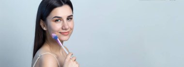 Açık renk bluzlu, beyaz arka planda yüz makyajı yapan genç bir kızın yakın çekimi. Güzel bir kadın yüzüne bir makyaj fırçası tutar ve gülümser..