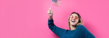 Stüdyonun pembe arka planında Amerikan bayrağı tutan mutlu genç kadın.